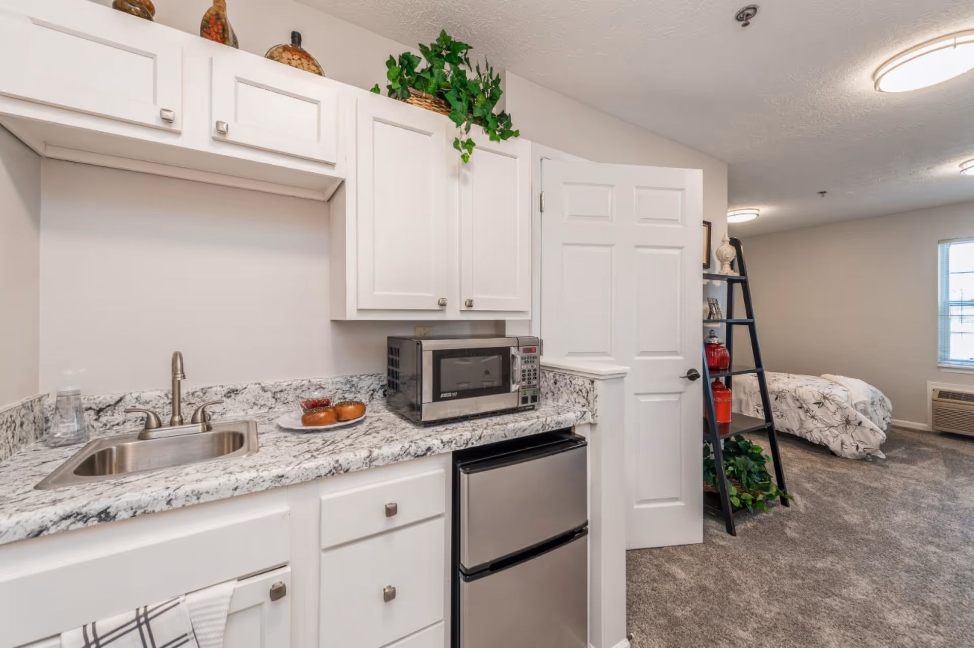 A small kitchenette area with white cabinets, a stainless steel sink, a microwave, and a mini refrigerator. The countertop has a plate with two bagels. In the background, there is a bedroom area with a bed, a window, and a black ladder-style shelf with decorative items and plants.
