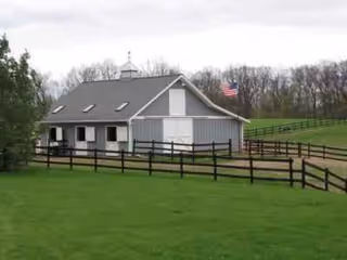 Gray barn-style assisted living building with white trim, fenced yard, and flag on green field.