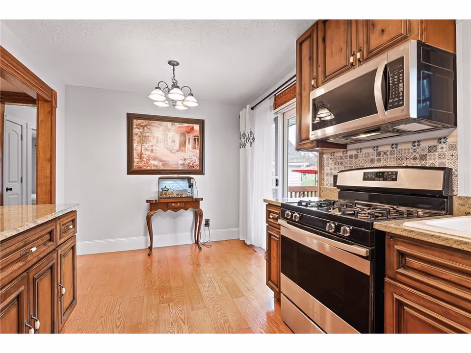 A kitchen area with wooden cabinets, a stainless steel gas stove and microwave, granite countertops, and a small wooden table with a fish tank on it. There is a framed painting on the wall above the table, a hanging light fixture on the ceiling, and a sliding glass door with white curtains leading outside.