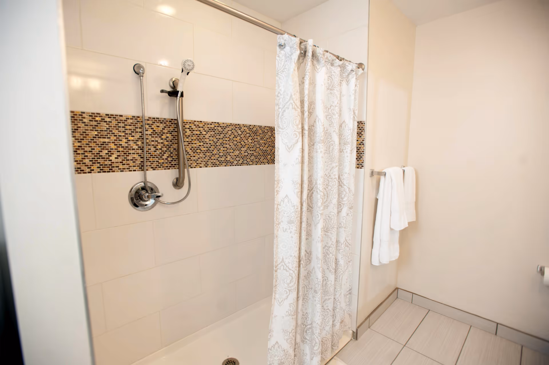 A clean bathroom shower area with a white and beige patterned shower curtain, a handheld showerhead mounted on a tiled wall with a horizontal strip of small brown mosaic tiles, and white towels hanging on a towel rack on the adjacent wall.