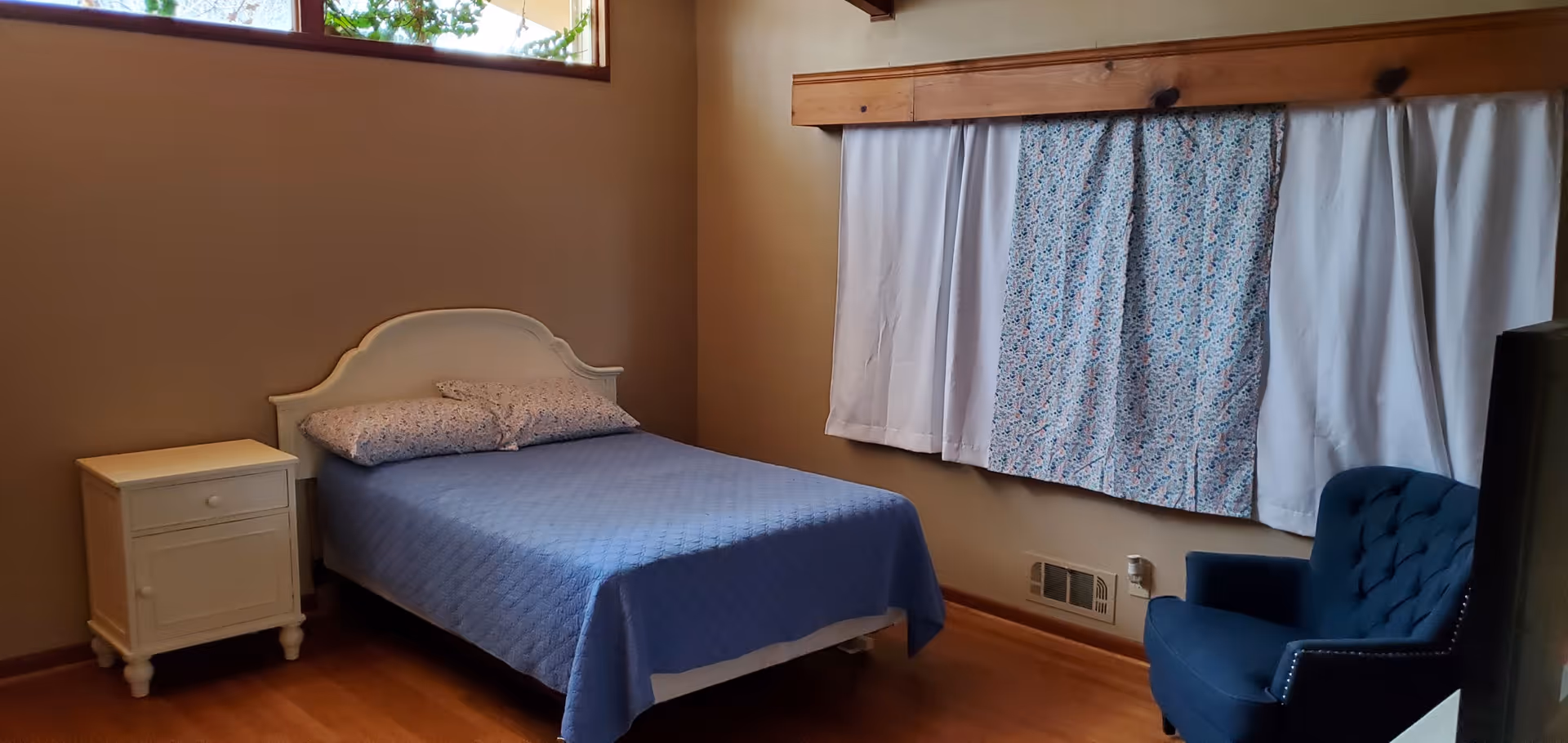 A simple bedroom with a white bed frame and blue quilted bedspread, two floral pillows, a white nightstand with a drawer and cabinet, a blue upholstered armchair, wooden floor, beige walls, and a window with white and floral curtains.