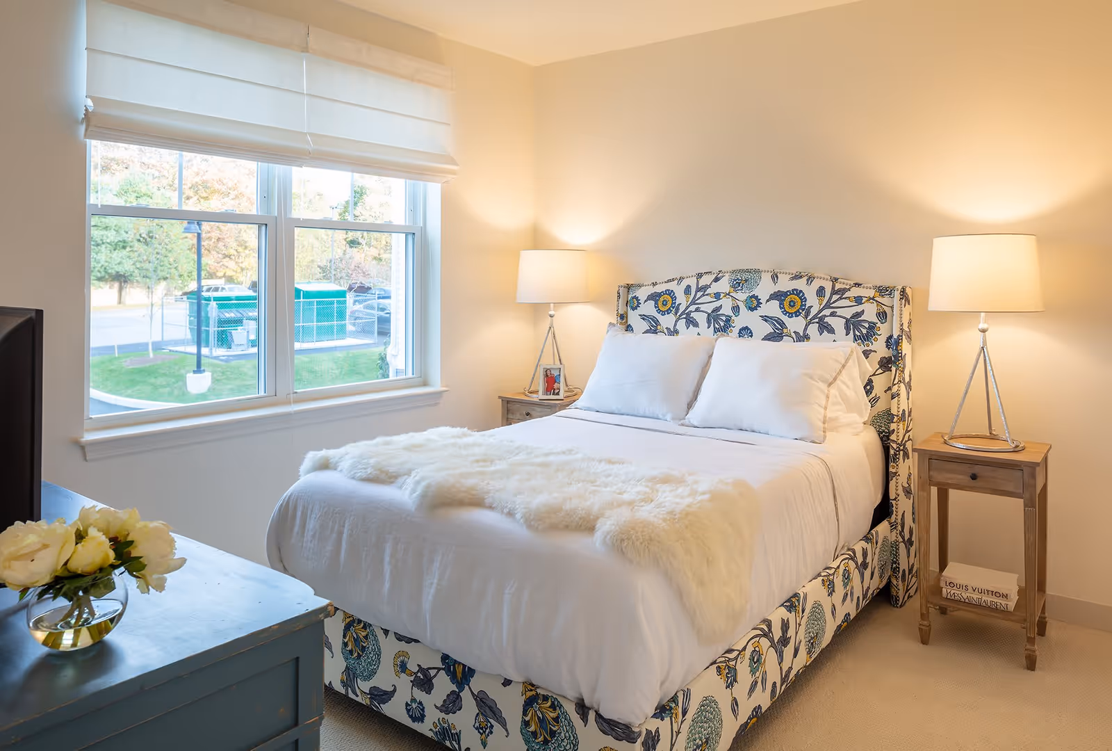 A bright and cozy bedroom with a large window showing an outdoor view. The bed has a floral patterned headboard and base, white bedding, and a fluffy white throw blanket. There are two bedside tables, each with a lamp, and a small framed photo on the left table. A blue dresser with a vase of white flowers is partially visible in the foreground.