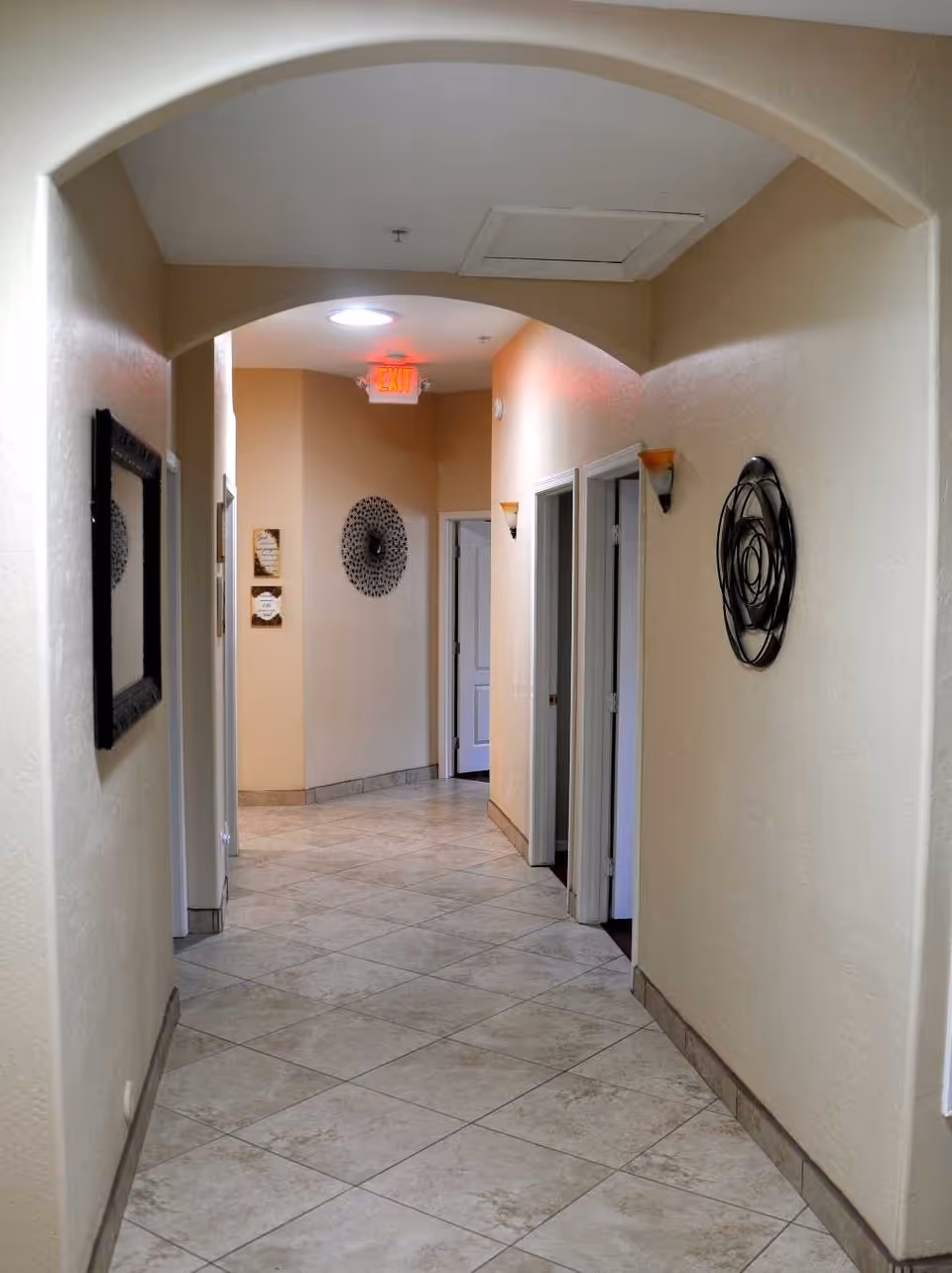 A clean, well-lit hallway with beige walls and tiled floor. The hallway features an arched ceiling, wall-mounted light fixtures, decorative wall art, and several open doorways leading to other rooms. An illuminated exit sign is visible at the end of the hallway.