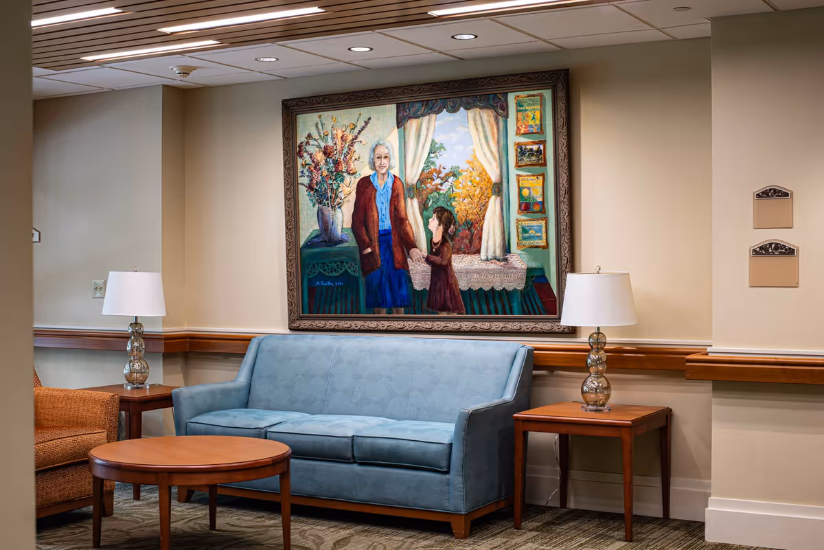 Comfortable seating area with a blue sofa, wooden side tables and lamps beneath a large framed painting on a hallway wall.