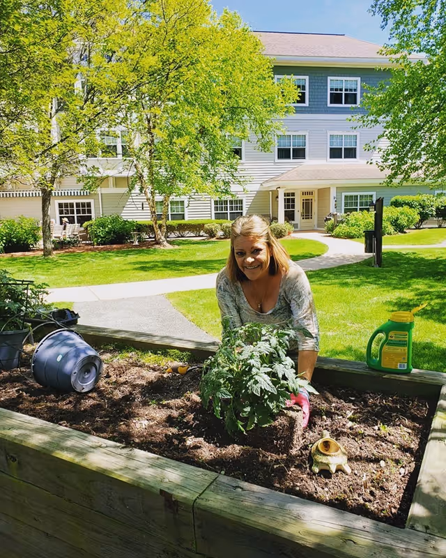 A woman is gardening in a raised wooden planter bed outside a senior living facility. She is planting a green leafy plant in the soil, wearing gardening gloves, and smiling at the camera. The background shows a well-maintained lawn, trees with green leaves, and a multi-story building with white siding and multiple windows under a clear blue sky.