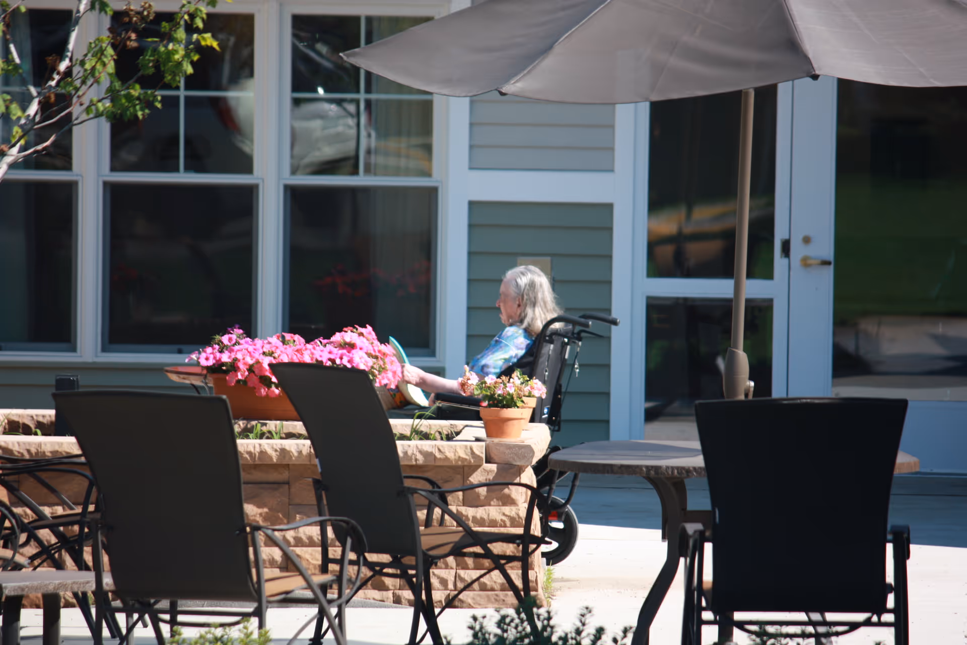 An elderly person sitting in a wheelchair outside on a patio near a flower bed with pink flowers. The patio has several chairs and tables with an umbrella, and the background shows a building with large windows and a door.