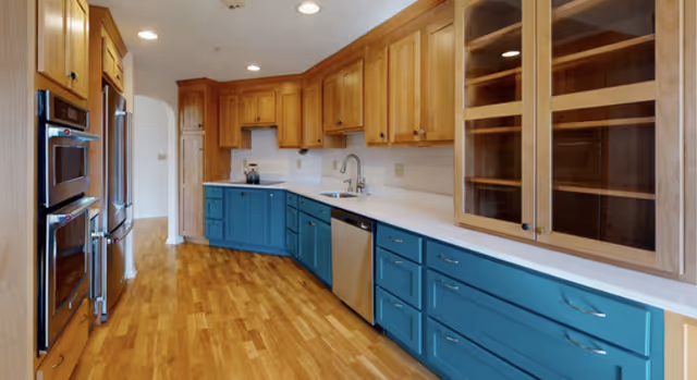 Spacious kitchen with natural wood upper cabinets, teal lower cabinets, stainless steel appliances, white countertops, and hardwood floors.