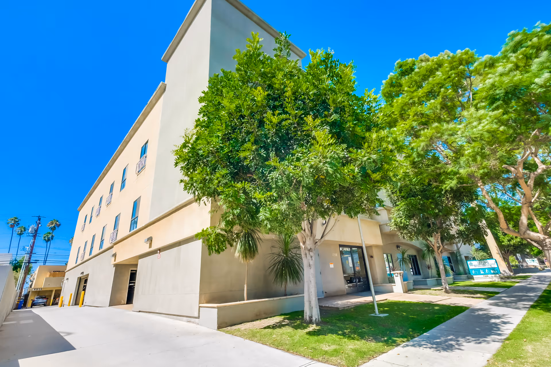 Exterior view of a multi-story beige building with several windows, surrounded by green trees and a sidewalk under a clear blue sky.