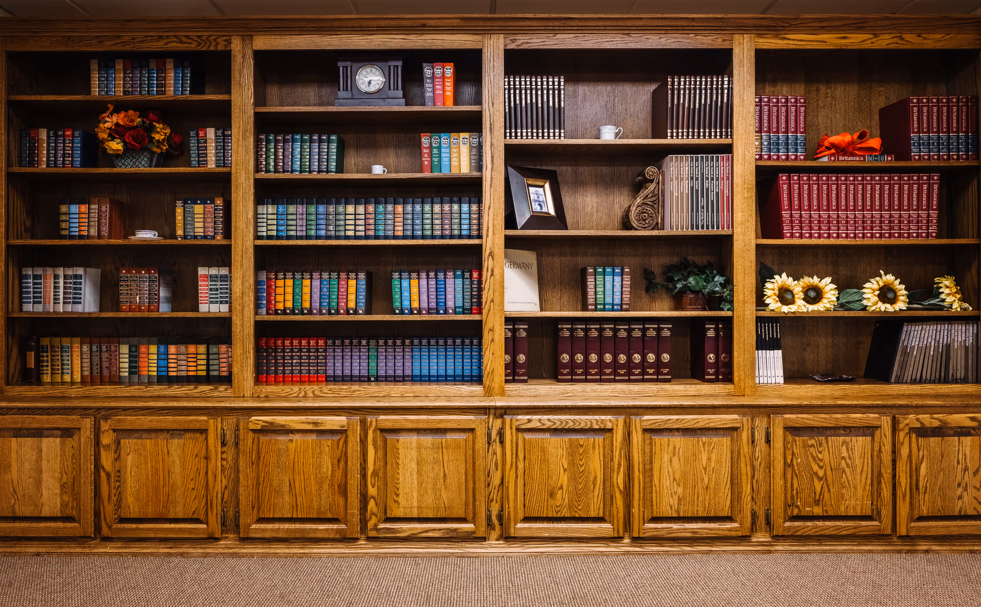 A large wooden bookshelf filled with numerous colorful books, decorative items including a clock, framed photo, artificial flowers, and plants. The bookshelf has multiple shelves and closed cabinets at the bottom, set against a carpeted floor.