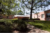 Outdoor view of Leroy Village Green Residential Health Care Facility showing a brick building surrounded by green grass, bushes, and trees with a paved walkway leading through the landscaped area.