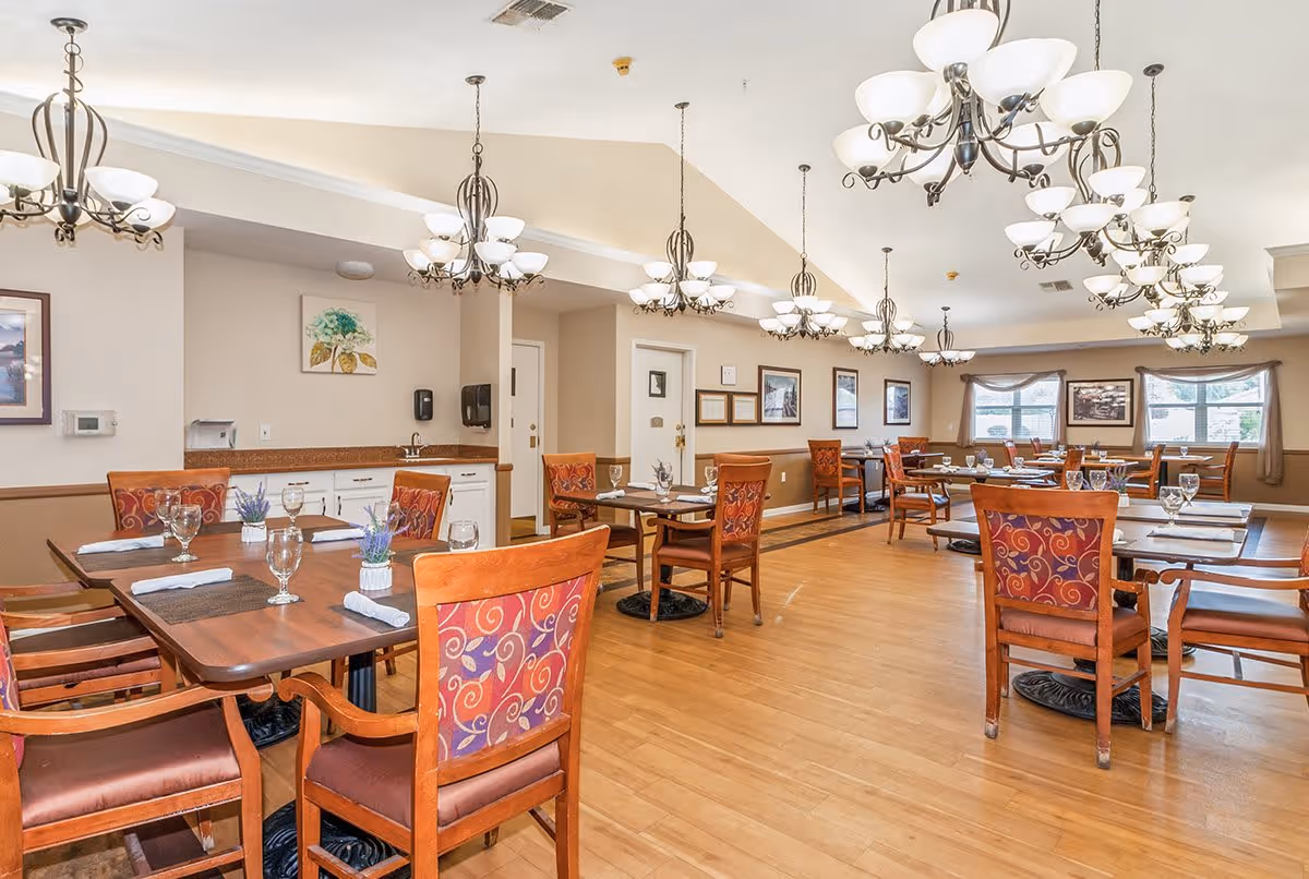 A spacious dining room with multiple wooden tables and chairs featuring red and purple patterned upholstery. Each table is set with placemats, napkins, and glassware. The room has wooden flooring, beige walls, several framed pictures, and large windows with curtains allowing natural light. Multiple elegant chandeliers hang from the ceiling providing warm lighting.