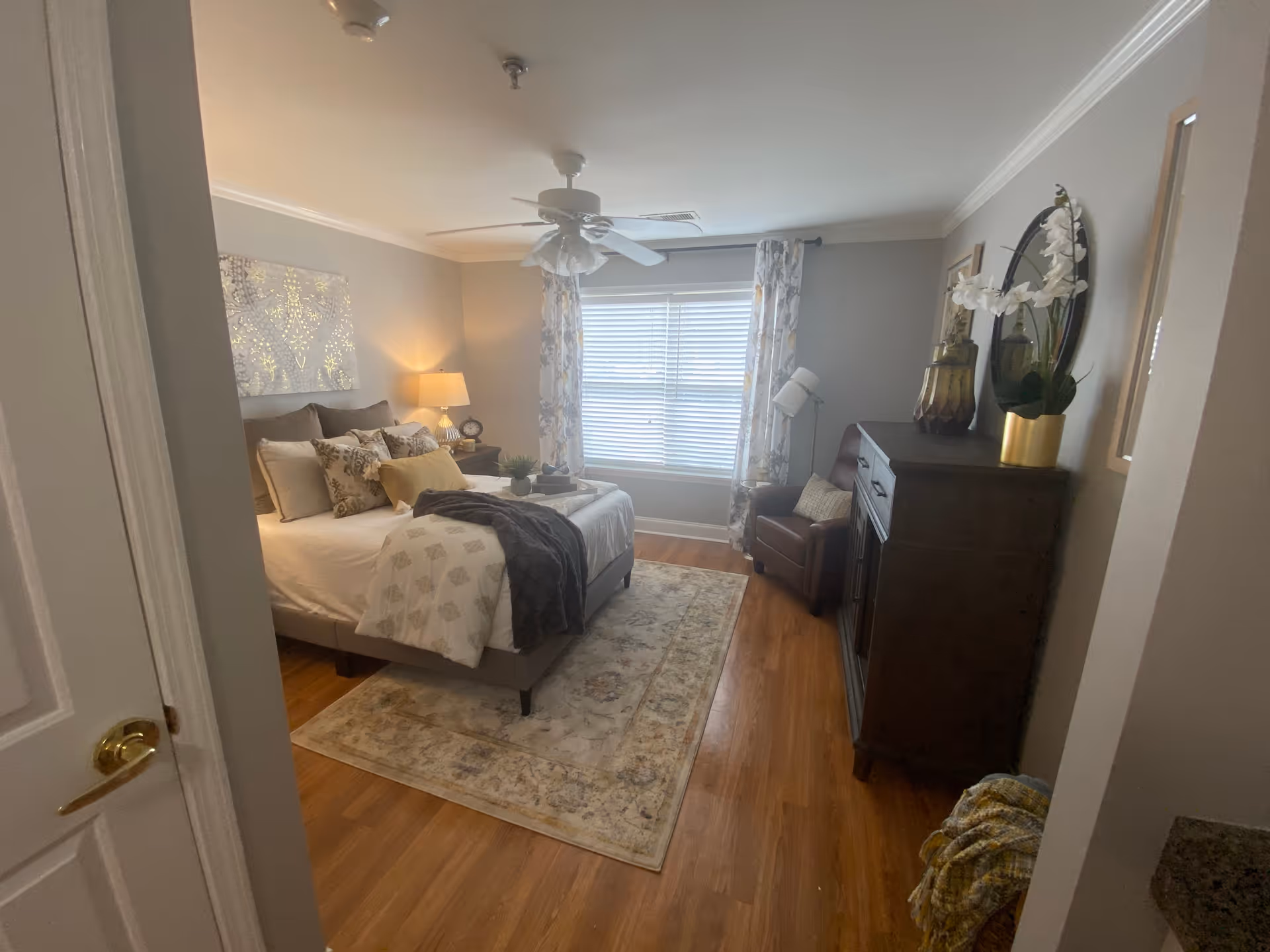 A cozy bedroom with a neatly made bed featuring multiple pillows and a throw blanket. There is a bedside table with a lamp and clock, a window with floral curtains, a ceiling fan, a patterned rug on wooden flooring, a dark wooden dresser with decorative items including a potted orchid, and a comfortable armchair with a cushion.