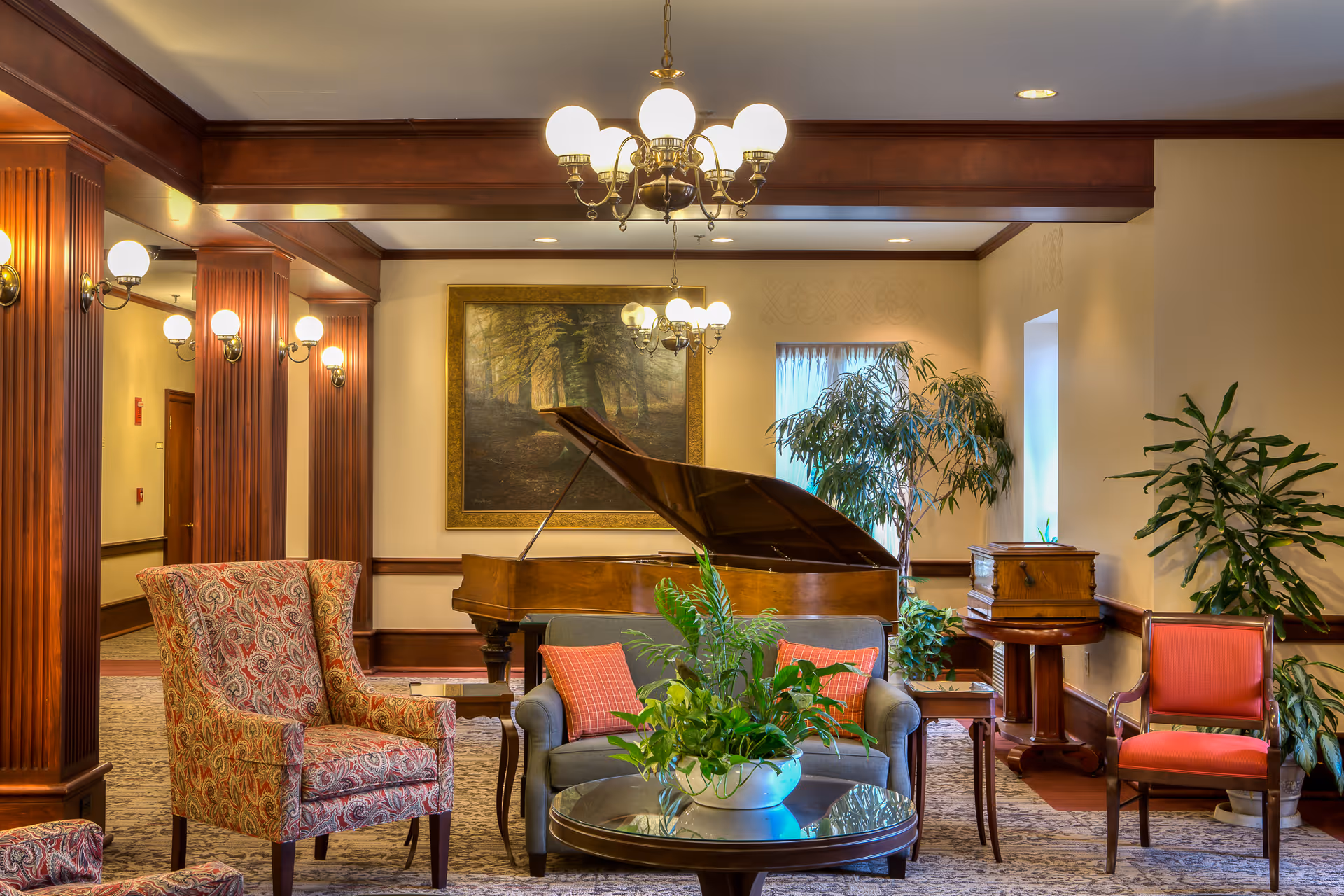 Elegant common room with a grand piano, upholstered chairs and sofa, plants, and chandeliers.