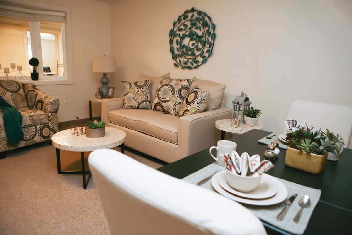 A cozy living room area with a beige loveseat adorned with patterned throw pillows, a matching armchair with a green throw blanket, two round side tables with decorative plants and a lantern, and a dining table set with white dishes, cups, and utensils. A decorative metal wall art piece hangs above the loveseat.