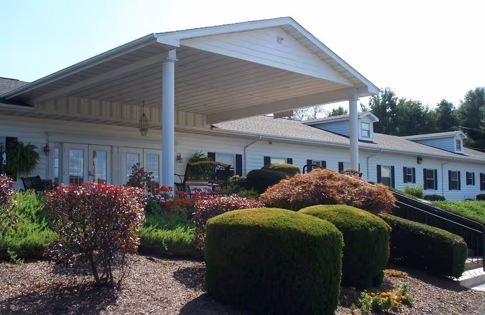 Exterior view of a single-story senior living facility with white siding, black shutters, and a covered porch area. The porch has seating and is surrounded by well-maintained bushes and flowering plants under a clear sky.