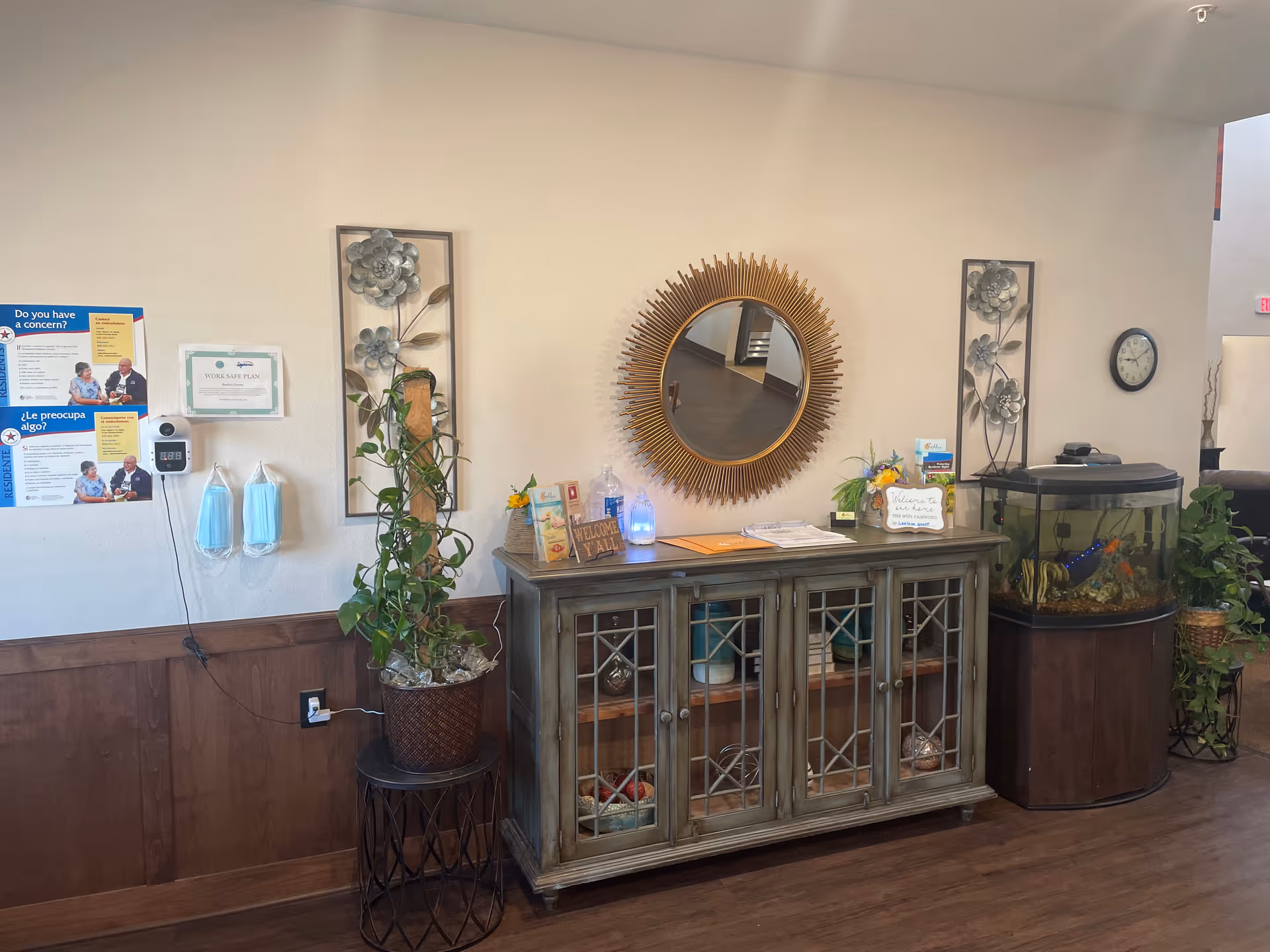 Interior view of a senior living facility reception or common area with a decorative wooden cabinet topped with plants, a round sunburst mirror, informational signs, and a fish tank. The wall has mounted metal floral decorations, a clock, and a hand sanitizer dispenser with face masks hanging below it.
