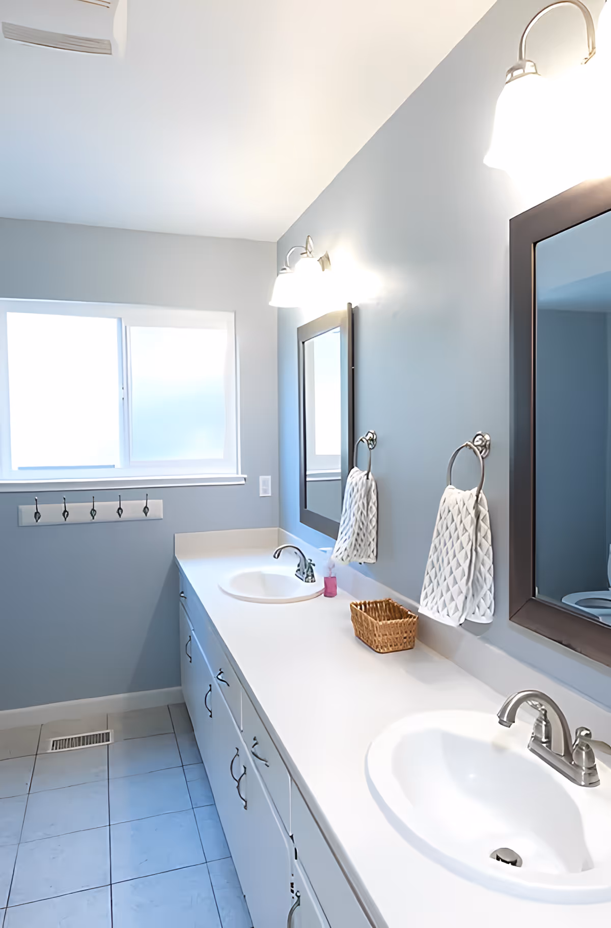 A clean bathroom with two white sinks set in a long white countertop. Above each sink is a rectangular mirror with a dark frame and a wall-mounted light fixture. A white towel hangs on a ring between the mirrors, and a small wicker basket and a pink soap dispenser are on the countertop. The walls are painted light blue, and there is a frosted window letting in natural light. The floor is tiled in a light color.