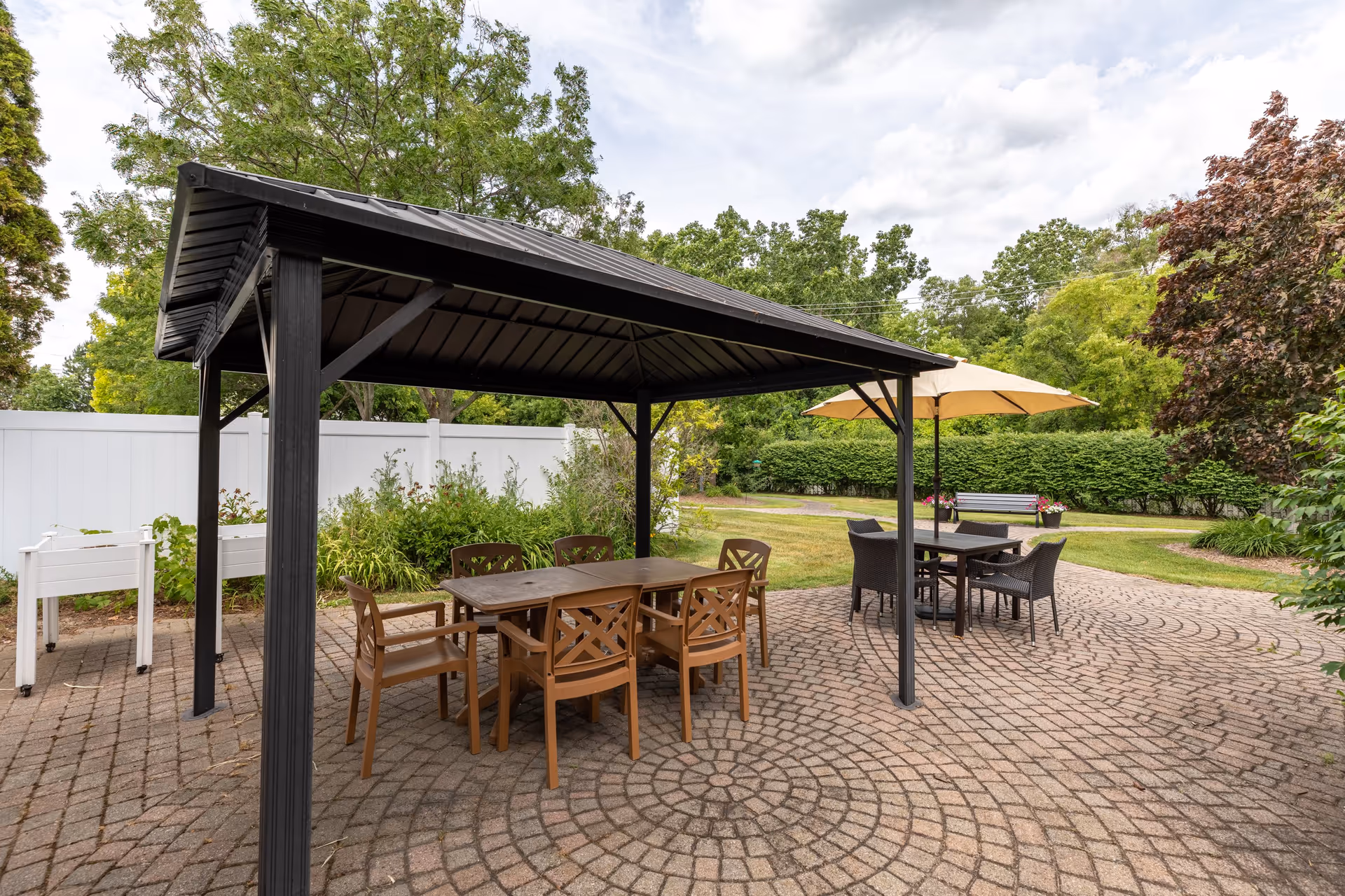 Outdoor patio area with a black metal gazebo covering a table and six brown chairs. Nearby is another table with four black chairs and a beige umbrella. The patio is paved with circular patterned bricks and surrounded by green trees, bushes, and a white fence.