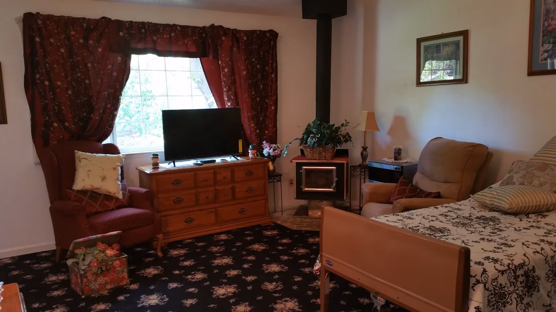 A cozy living space with a floral carpet, a red armchair with pillows, a wooden dresser with a flat-screen TV on top, a wood stove with a plant on it, a beige recliner chair, a bed with patterned bedding, and framed pictures on the wall.