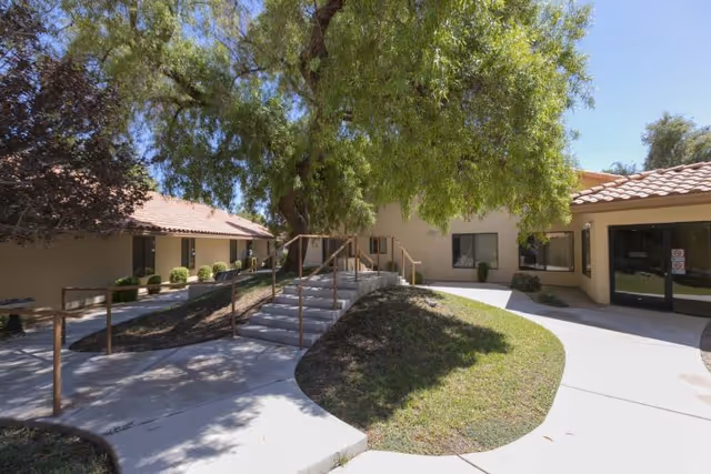 Outdoor view of a senior living facility courtyard with a concrete pathway, stairs with handrails, green grass, and large trees providing shade. The building has beige walls and red-tiled roofs with windows and glass doors visible.