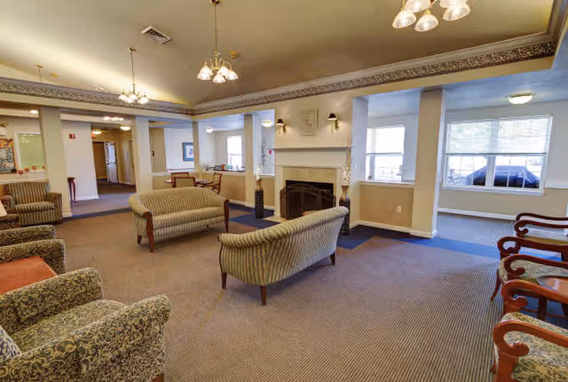 A spacious living room area in The Addison of Logan Place featuring multiple upholstered armchairs and sofas arranged around a central fireplace. The room has beige walls, carpeted flooring, and several windows allowing natural light. There are ceiling light fixtures and a decorative crown molding along the ceiling edges.
