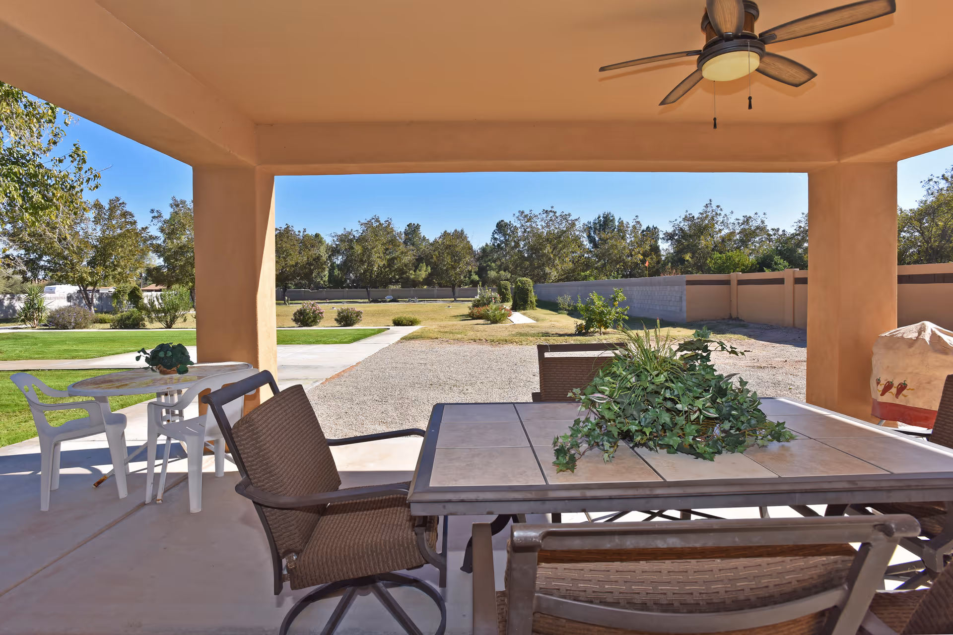 Covered patio with a dining table, chairs, ceiling fan and potted plant overlooking a lawn and trees.
