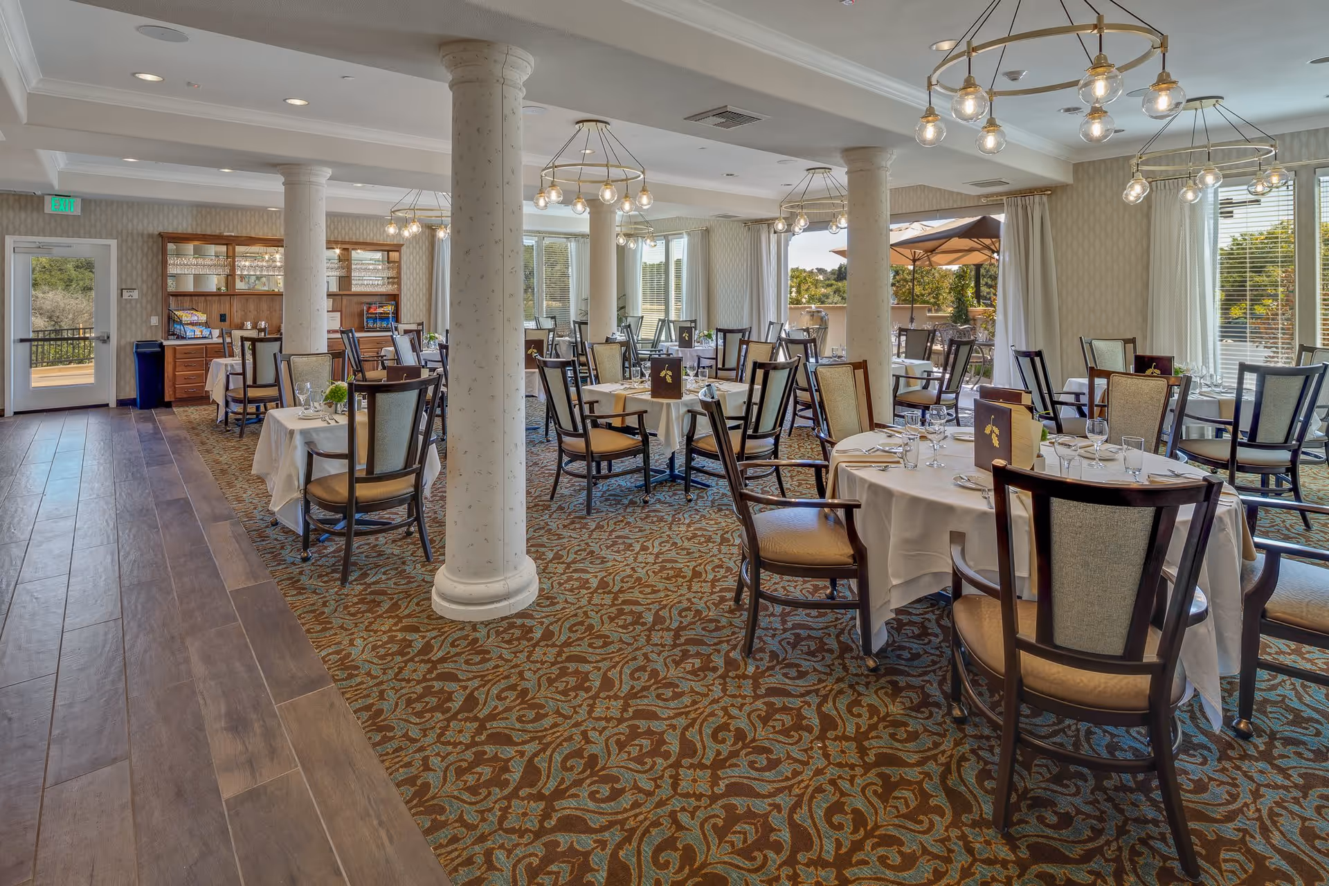 A spacious dining room with multiple round and rectangular tables covered with white tablecloths, set with glassware and menus. The room features patterned carpet, wooden chairs with cushioned seats, white pillars, large windows with white curtains, and modern hanging light fixtures. Outside the windows, there is a patio area with umbrellas and greenery.