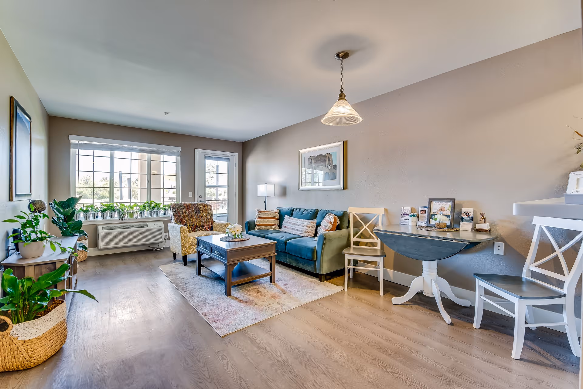 A bright and cozy living room with a teal sofa, a patterned armchair, a wooden coffee table on a rug, and a small round dining table with chairs. The room has large windows with white blinds, several potted plants, light wood flooring, and neutral-colored walls with framed artwork.