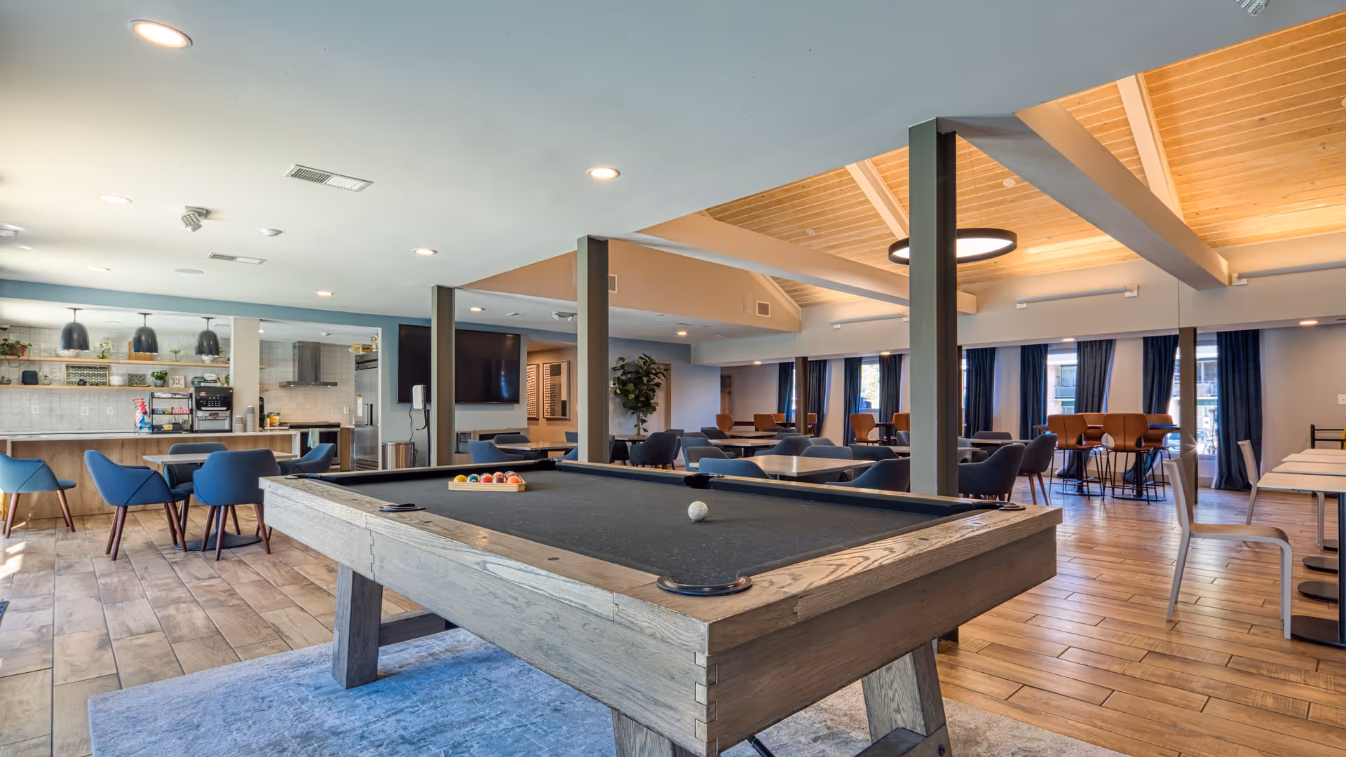 A spacious community room featuring a pool table with balls arranged for a game, multiple seating areas with tables and chairs, a kitchenette with pendant lights, and large windows with dark curtains allowing natural light to enter. The ceiling has wooden beams and recessed lighting.
