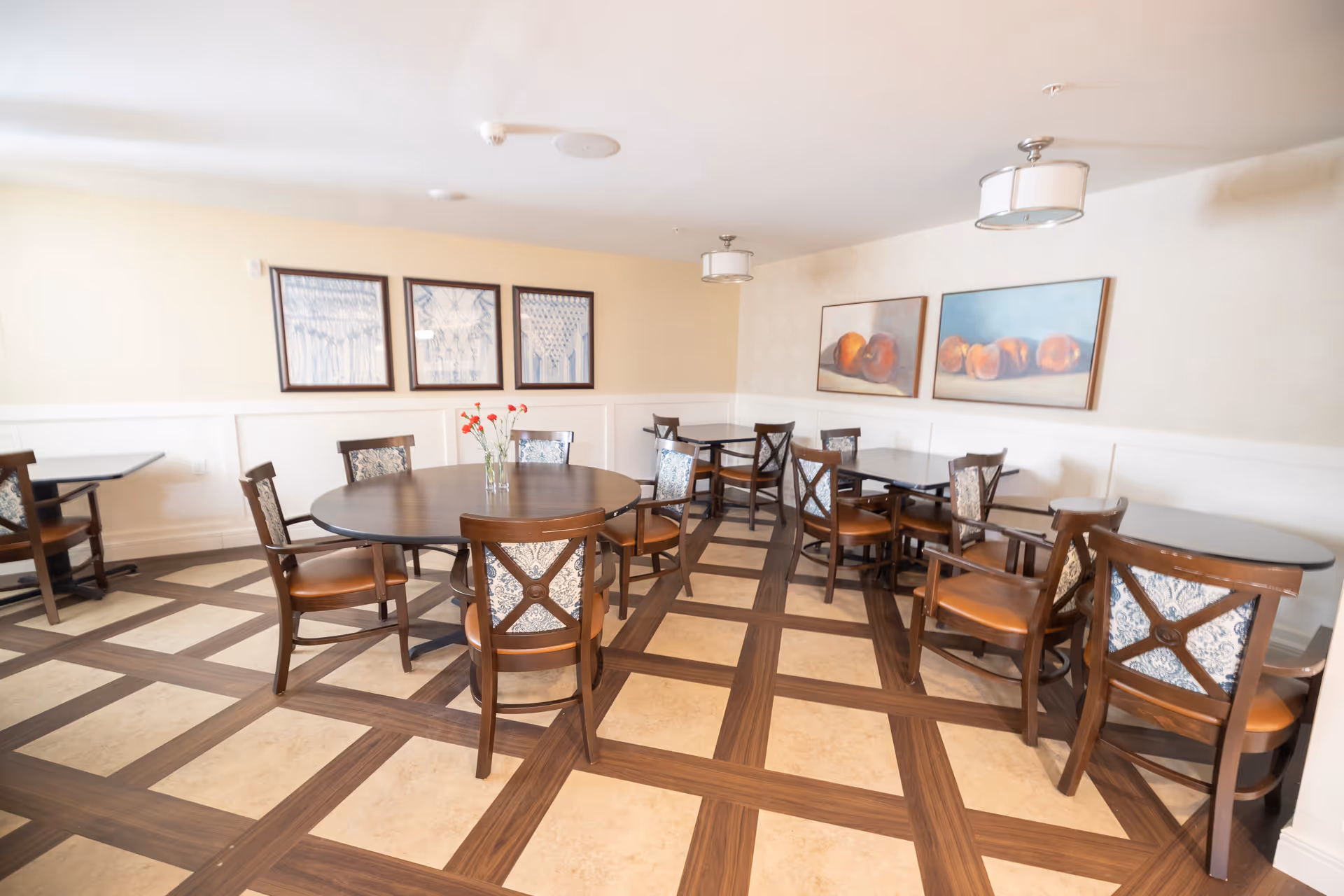 A bright dining room with several wooden tables and chairs arranged neatly. The floor has a checkered pattern with wood and beige tiles. The walls are decorated with framed artwork, including images of peaches and abstract designs. A vase with red flowers is placed on the central round table.