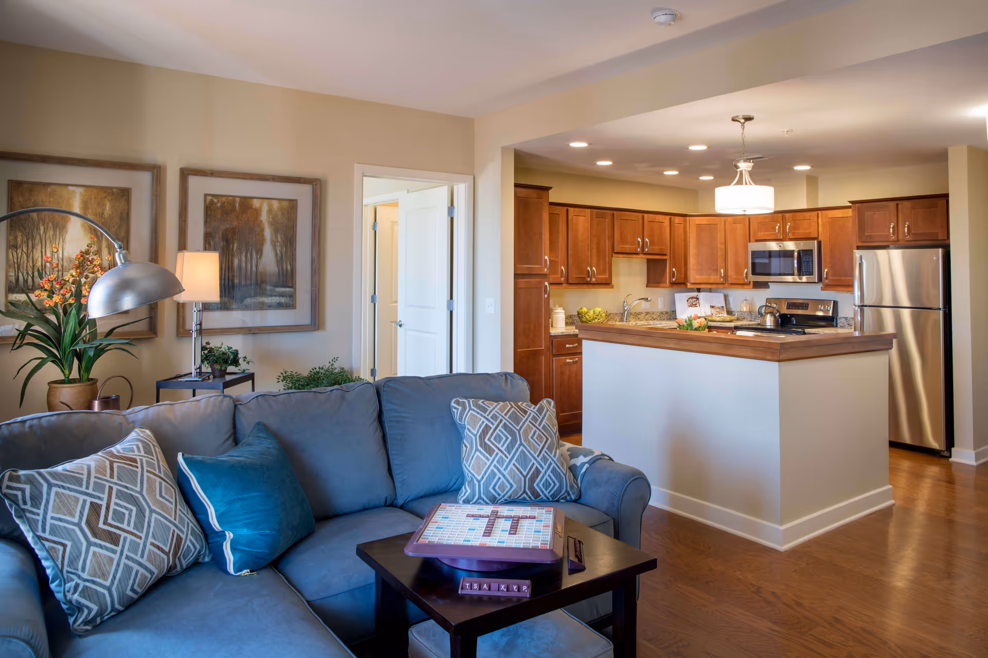 A cozy living room area with a blue-gray sofa adorned with patterned and solid pillows, a small dark wooden coffee table with a Scrabble game on it, a floor lamp, and two framed paintings on the wall. The living room opens into a kitchen with wooden cabinets, granite countertops, stainless steel appliances, and a hanging light fixture above the kitchen island.