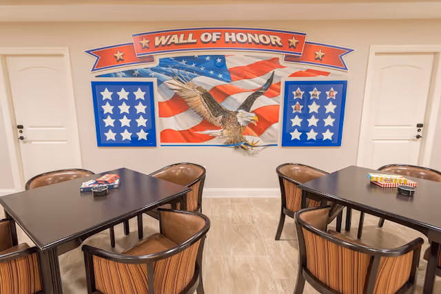 A room with two dark wooden tables surrounded by six brown cushioned chairs. On the wall behind the tables is a patriotic display titled 'Wall of Honor' featuring an American flag, a bald eagle, and two blue boards with white stars.