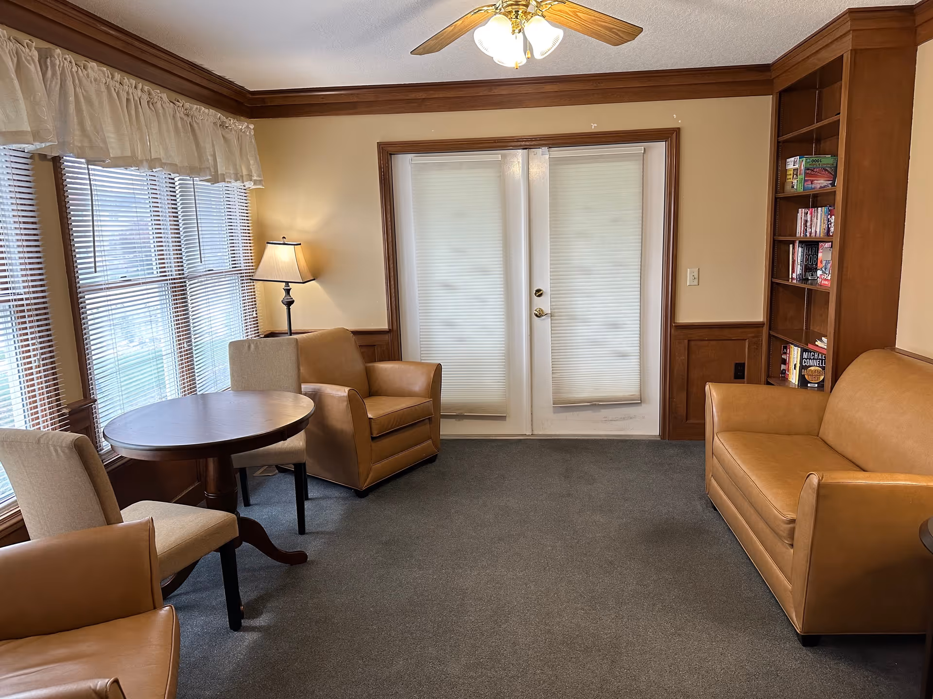 A cozy living room with beige walls and wood trim, featuring a ceiling fan with lights, a round wooden table with two beige upholstered chairs, two tan leather armchairs, a tan leather sofa, a floor lamp, and a wooden bookshelf filled with books. The room has large windows with blinds and sheer curtains, and white double doors with blinds.