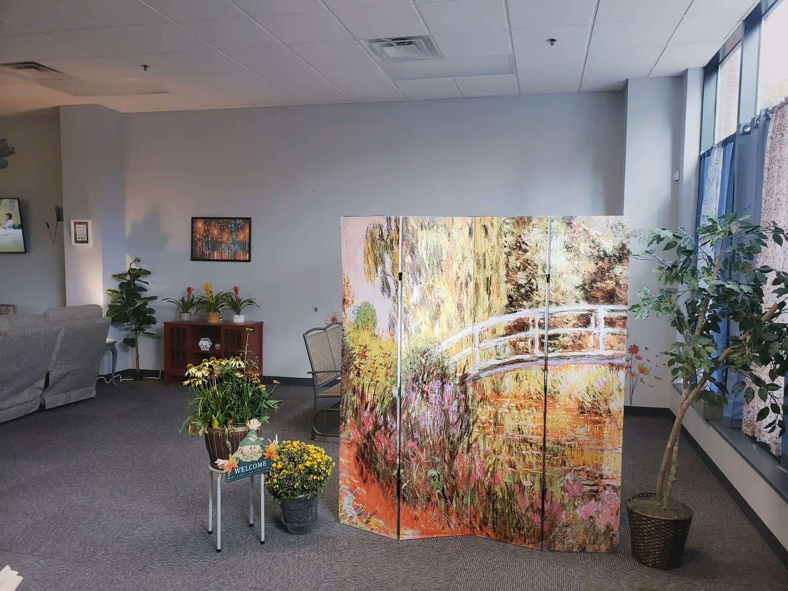 A bright common room with a decorative painted folding screen, several potted plants, seating, and large windows.