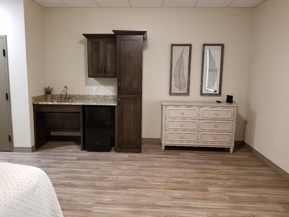Interior view of a room with a small kitchenette area featuring a granite countertop, a sink, dark wooden cabinets, and a mini refrigerator. Next to the kitchenette is a white dresser with six drawers and two framed leaf artworks hanging above it. The floor is wood laminate, and the walls are painted light beige.