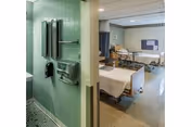 A rehab facility patient room with two hospital beds, privacy curtains, medical furnishings, and a green-tiled sink area in the foreground.