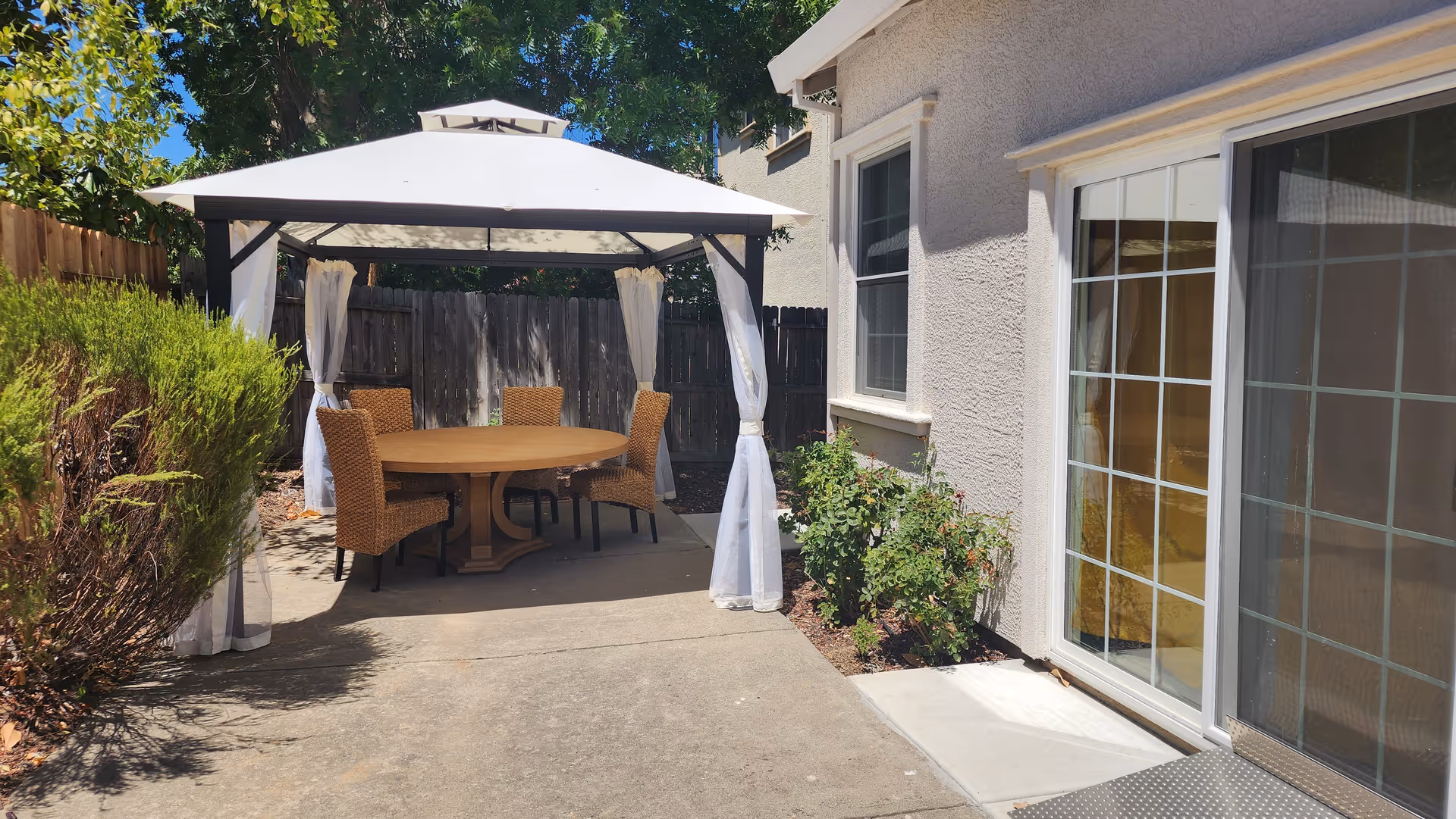 Outdoor patio area with a white canopy tent covering a round wooden table surrounded by four wicker chairs. The patio is adjacent to a beige building with a sliding glass door and a window. There are bushes and plants along the side of the building and a wooden fence in the background.