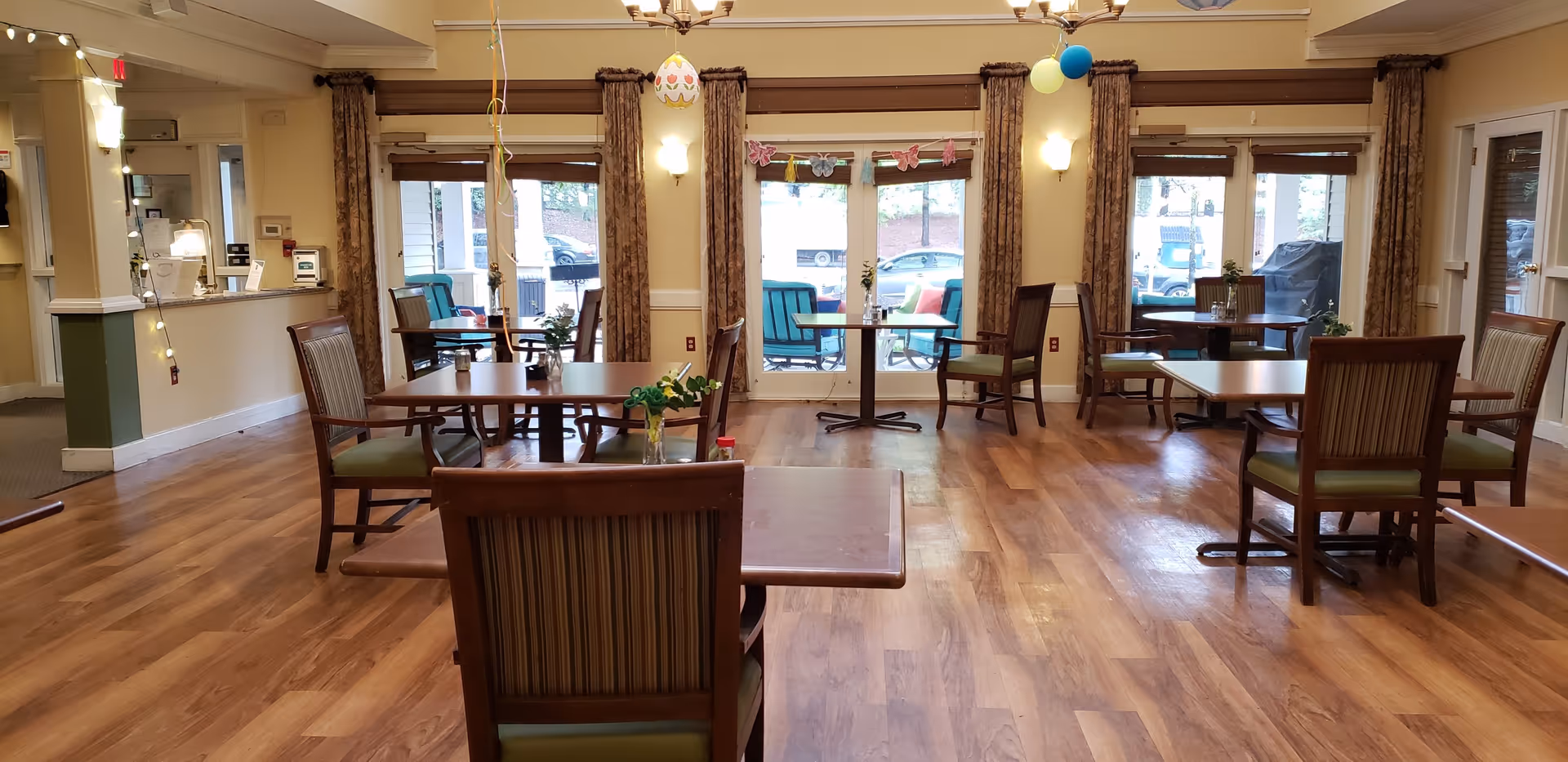 A dining room in a senior living facility with wooden tables and chairs arranged neatly. The room has large windows with brown curtains, letting in natural light. There are some decorative hanging ornaments and small flower vases on the tables. Outside the windows, patio furniture is visible.