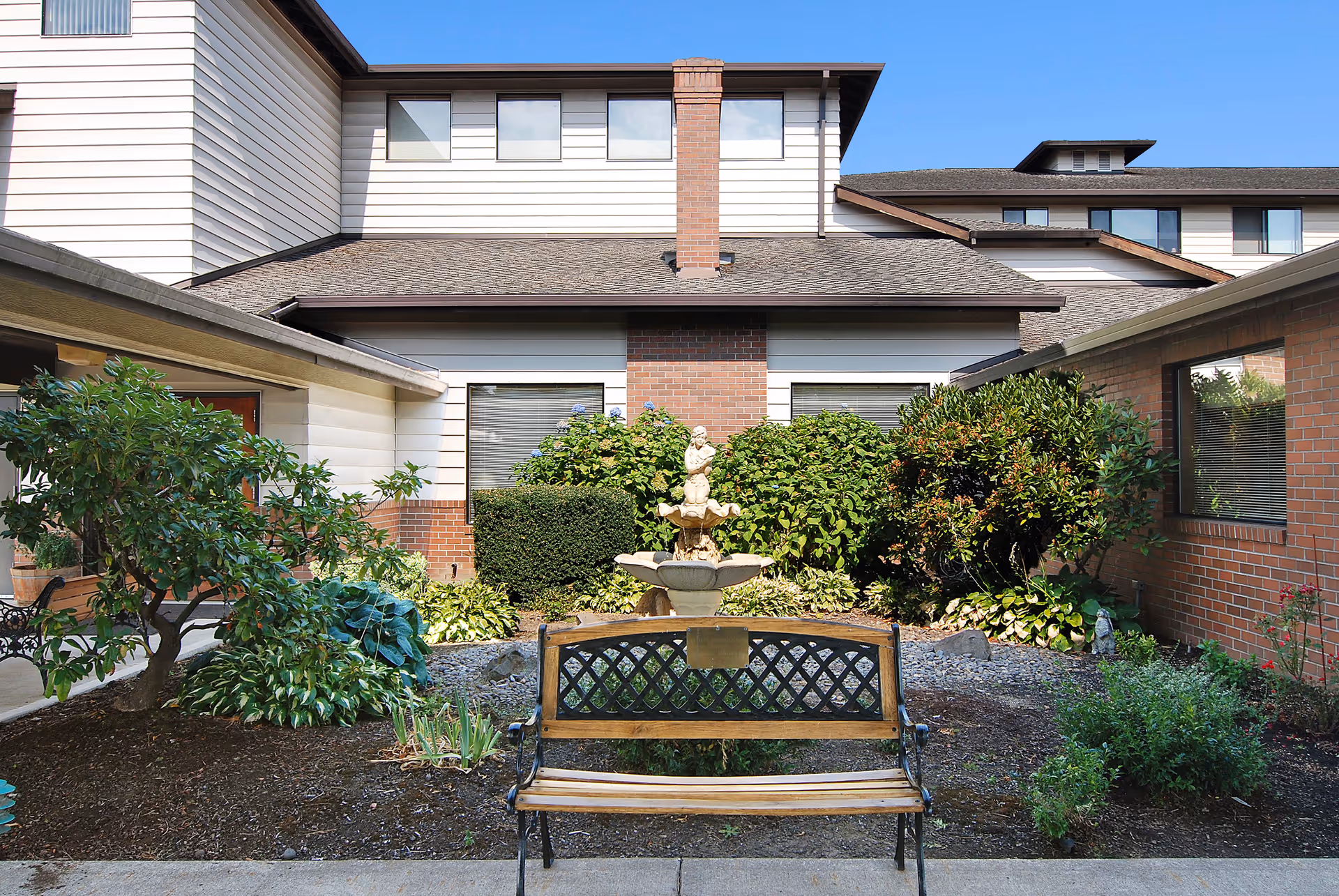 Courtyard with a bench and a small fountain surrounded by shrubs in front of a multi-story brick and siding building.