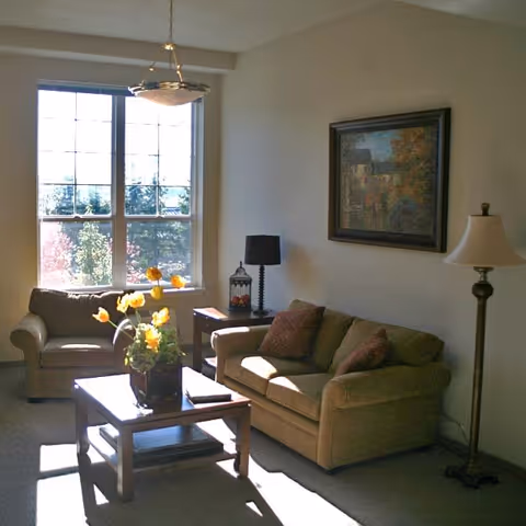 A sunlit living room with a large window showing trees outside. The room contains a beige loveseat and armchair with brown cushions, a wooden coffee table with a vase of yellow flowers, a side table with a decorative lantern and a black lamp, a floor lamp, and a framed painting on the wall.