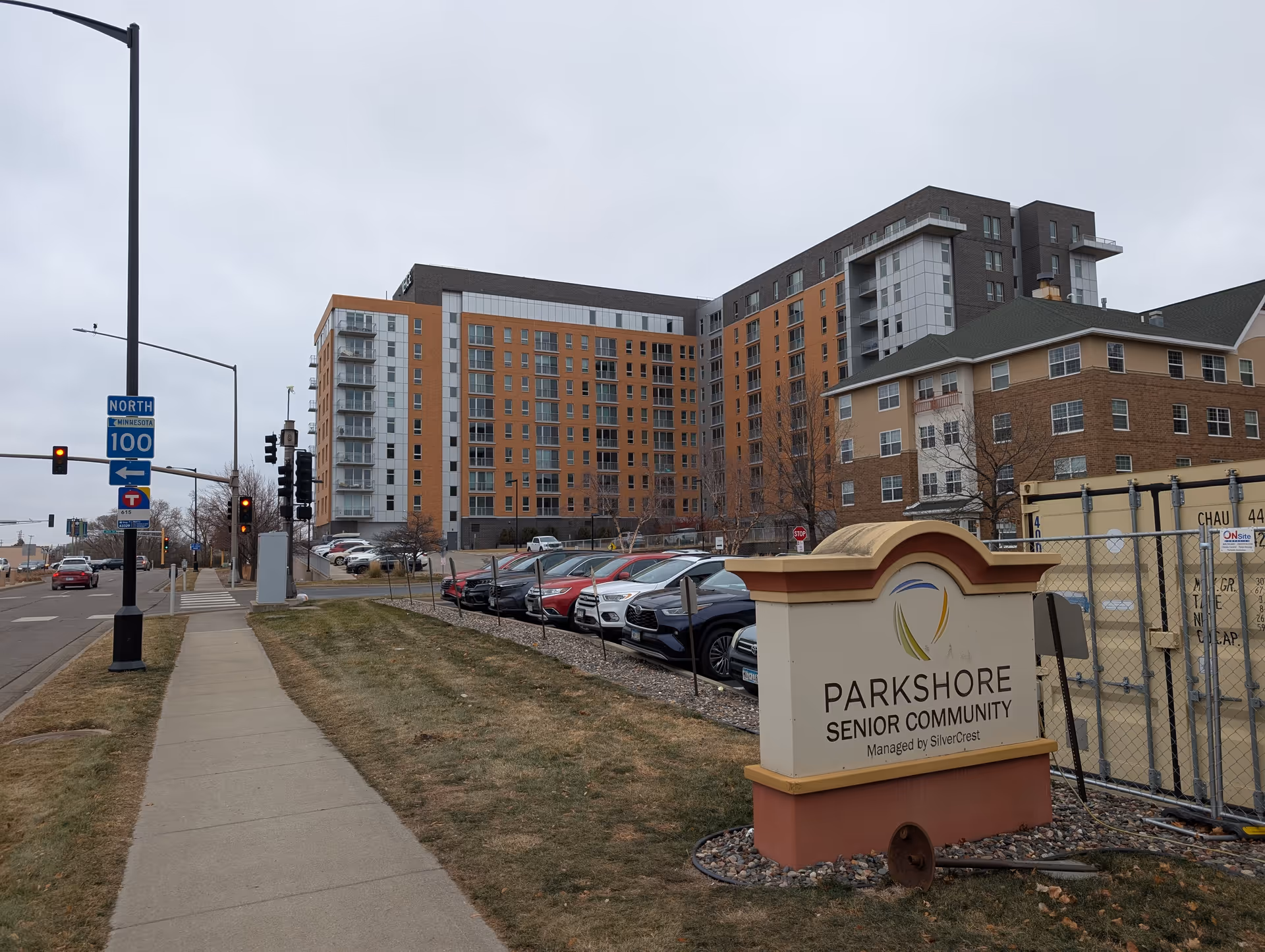 Street view showing the Parkshore Senior Community sign in the foreground, with the multi-story senior living building, parked cars, sidewalk and street signs under an overcast sky.