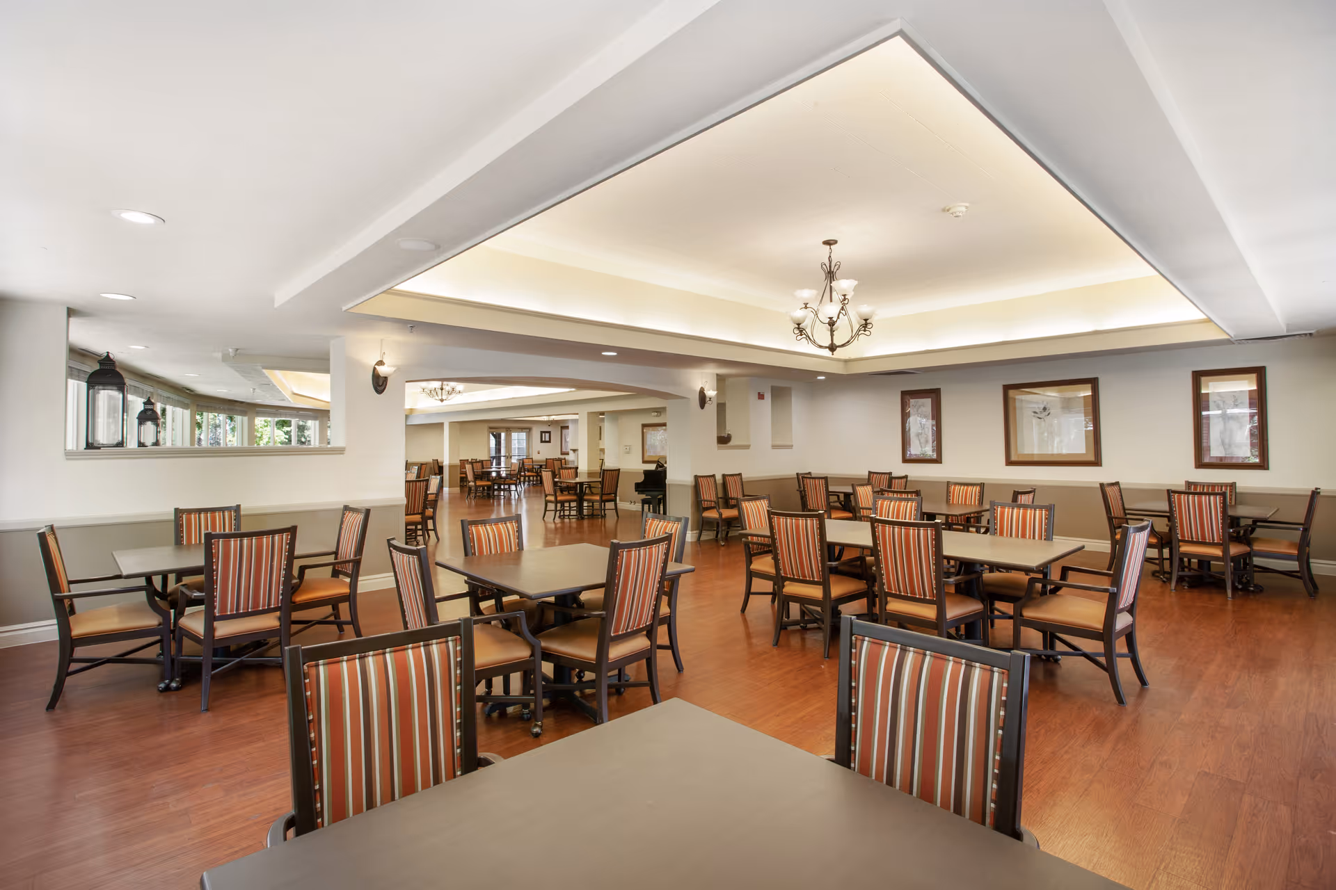 A spacious dining room with multiple tables and chairs arranged neatly. The chairs have striped upholstery and the room features wood flooring, framed artwork on the walls, and a chandelier hanging from a recessed ceiling with soft lighting.