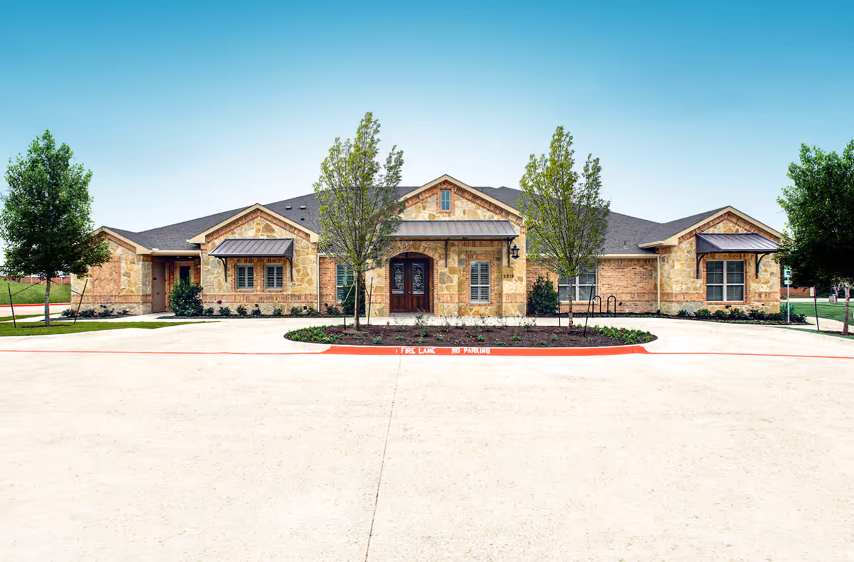 Single-story stone and brick senior living building with a central entrance, small trees, and a circular driveway.