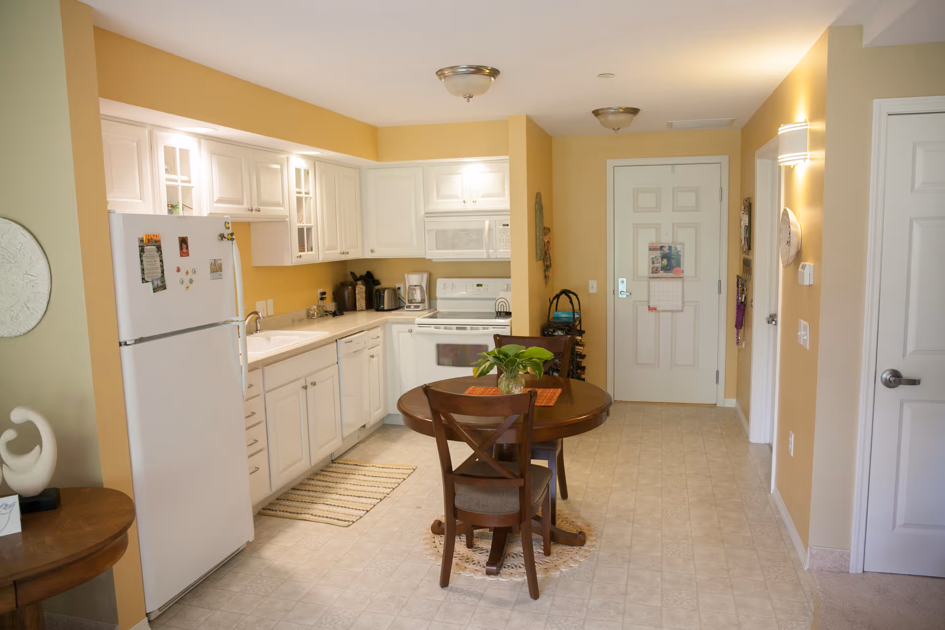 A bright kitchen area with white cabinets, a white refrigerator, stove, microwave, and dishwasher. A round wooden dining table with two chairs is placed in the center, with a small plant on top. The walls are painted yellow, and there is a door at the far end of the room. The floor is tiled in a light color.