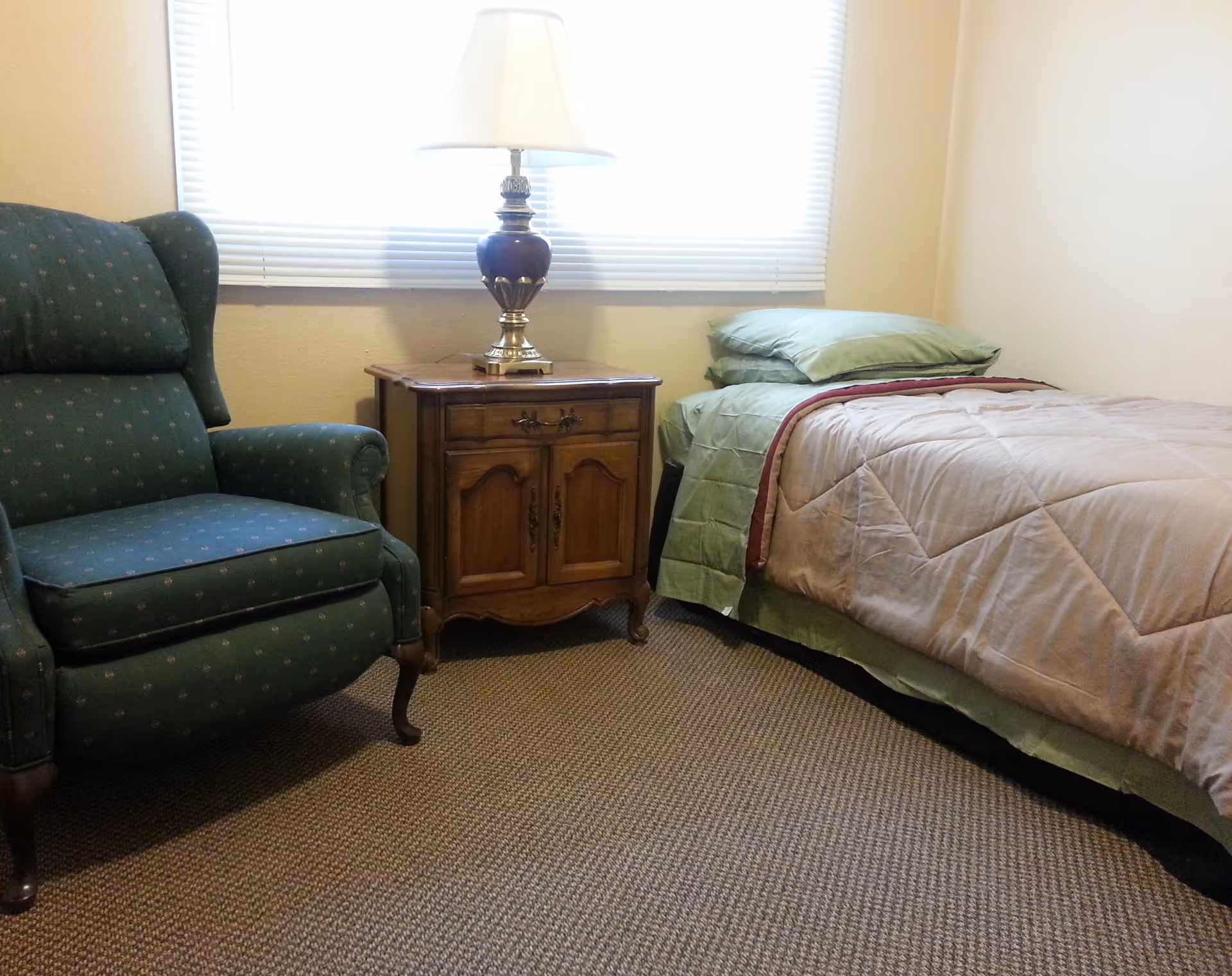 A small bedroom with a single bed made with green and beige bedding, a wooden nightstand with a decorative lamp, and a green upholstered armchair. A window with blinds is behind the nightstand.