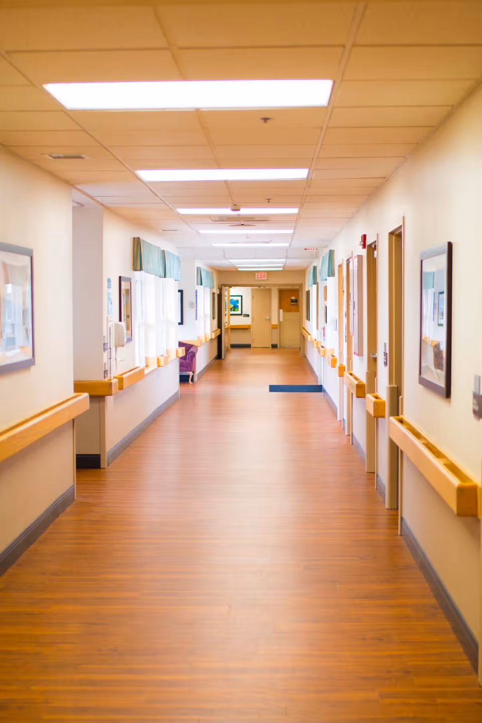 A long, well-lit hallway in a senior living facility with wooden floors, handrails on both sides, framed pictures on the walls, and windows with green valances letting in natural light. Doors line the right side of the hallway, and there is an exit door at the far end.