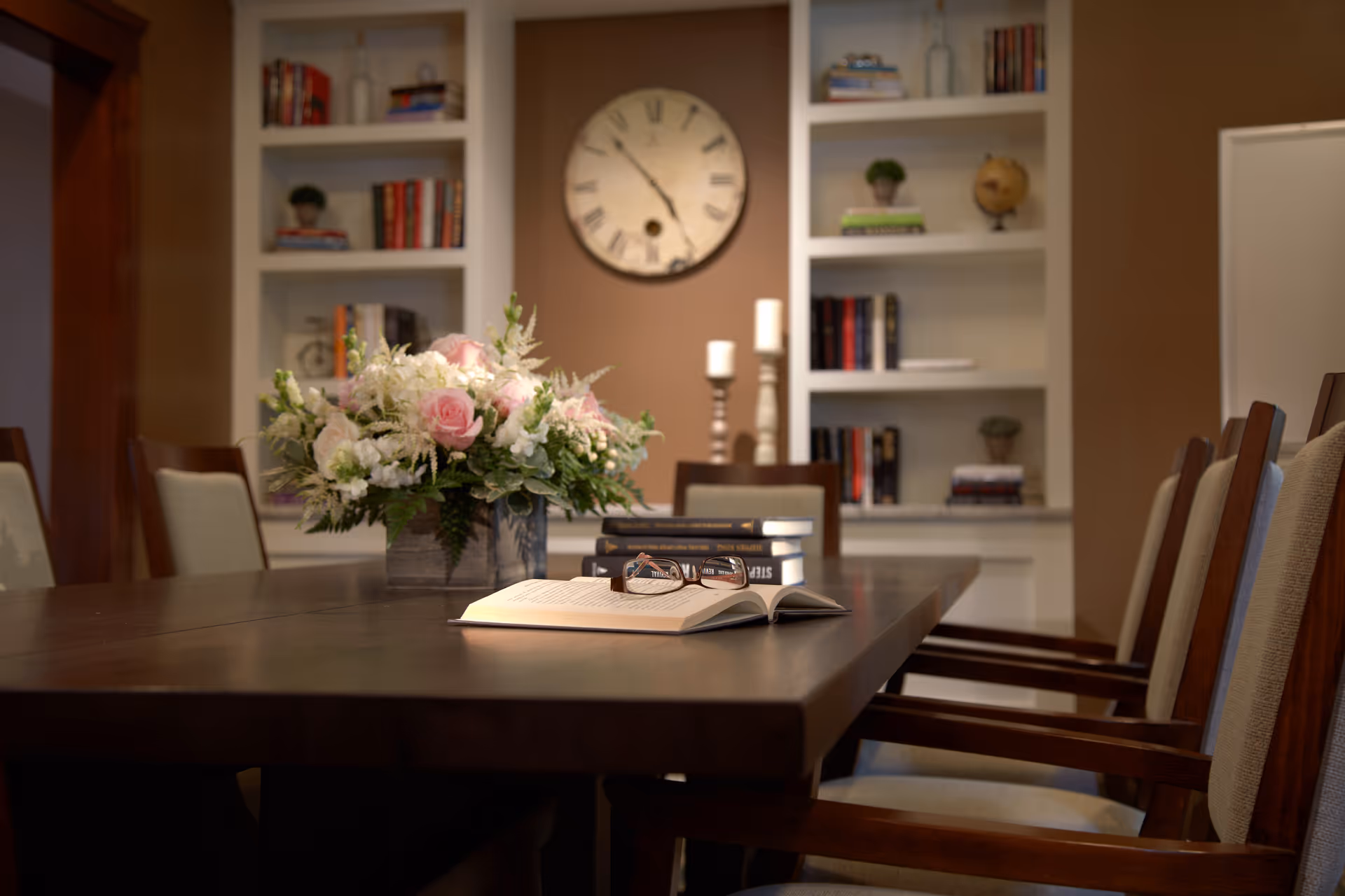 A dining room with a dark wooden table surrounded by cushioned chairs. On the table, there is a bouquet of flowers, an open book with reading glasses on top, and a stack of books. In the background, there are built-in white shelves filled with books and decorative items, a large wall clock, and two candle holders.
