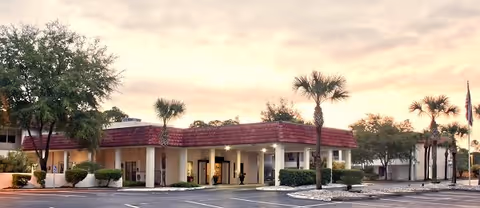 Exterior view of a single-story nursing and rehabilitation facility building with a red-tiled roof, palm trees, and a parking lot in front during sunset.