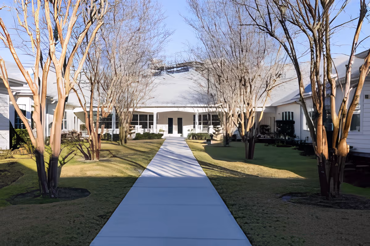 A paved walkway through a landscaped courtyard lined with trees leading to the entrance of a single-story building.