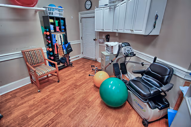 Small exercise/therapy room with a recumbent bike, two large stability balls, a rack of weights and equipment, a chair, and wall cabinets.